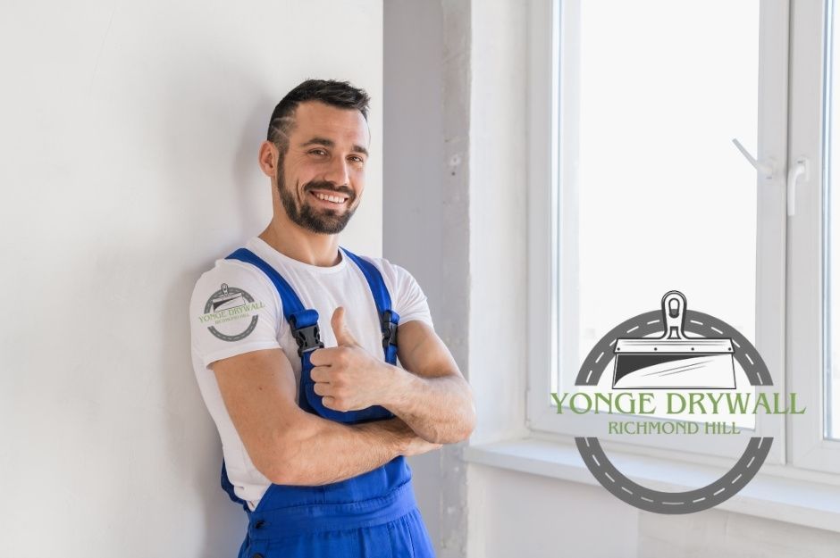 A drywall repair company worker wearing blue overalls and a white t-shirt stands beside a freshly finished commercial wall, smiling confidently and giving a thumbs-up gesture. Bright natural light enters through a nearby window, showing a clean, smooth surface near Emery Hill Blvd, and Stonebridge Dr, Markham, ON.