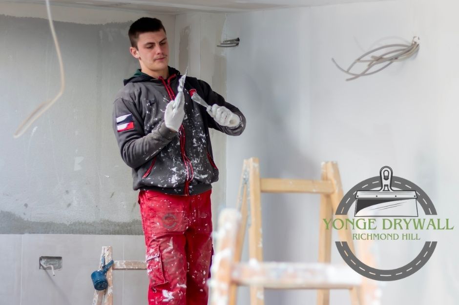 A drywall repair company worker is inside a commercial building, standing on a ladder while wearing red pants and a gray hoodie splattered with white plaster. He is holding two drywall knives, carefully scraping one against the other as he prepares for wall finishing near Cottingham Crescent, and Tilbury St, Oshawa, ON.