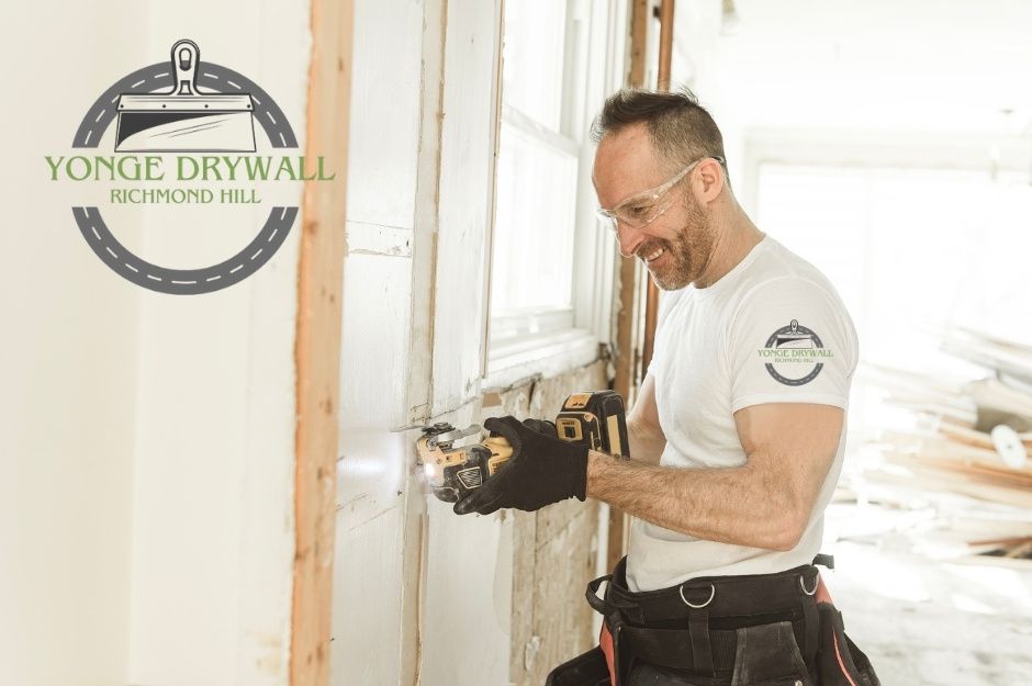 A drywall repair contractor wearing safety goggles, black gloves, and a white t-shirt uses a yellow oscillating multi-tool to cut into drywall near a window during a residential renovation. Sunlight fills the workspace as he focuses carefully. Located near Timberlane Dr, and Ravenswood Dr, Brampton, ON.