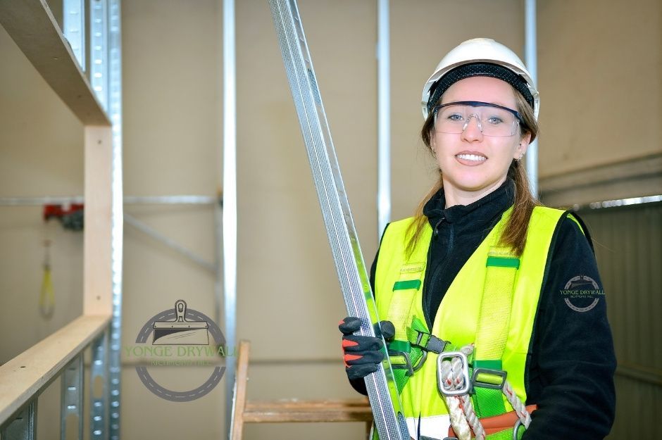 A drywall repair contractor in a bright yellow safety vest and hard hat smiles while holding metal framing studs in a commercial project with metal framing near Brock Concession Rd, and Trans - Canada Hwy, Brock, ON.
