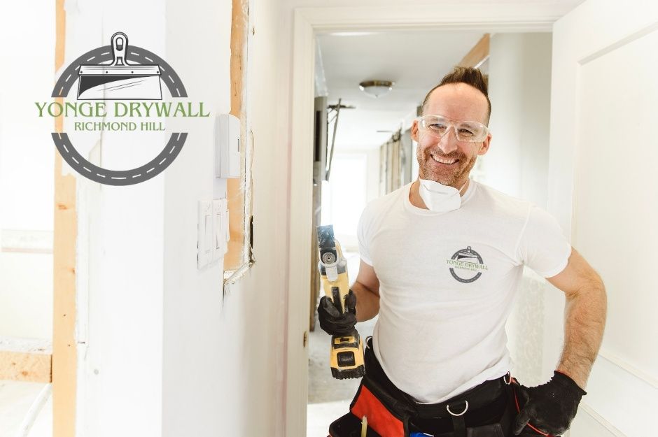 A drywall repair contractor wearing safety goggles, black gloves, and a white t-shirt smiles while holding a yellow oscillating multi-tool inside a hallway under renovation. A dust mask hangs around his neck, and tools fill his belt. A residential project near Balmoral Heights, and Testa St, East Gwillimbury, ON.