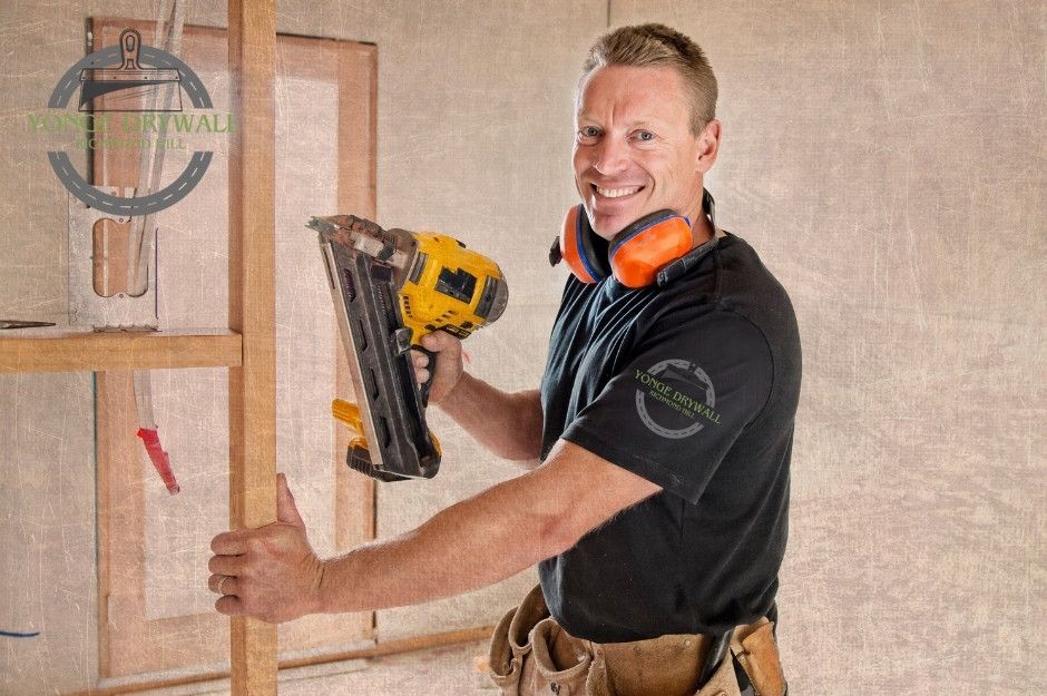 A drywall repair contractor smiles while working on a residential project with unfinished walls. He holds a yellow power nail gun in his right hand, while his left hand rests on a wooden frame. He wears protective orange ear muffs, a black t-shirt, and a tool belt near Vista Dr, and Turney Dr, Mississauga, ON.