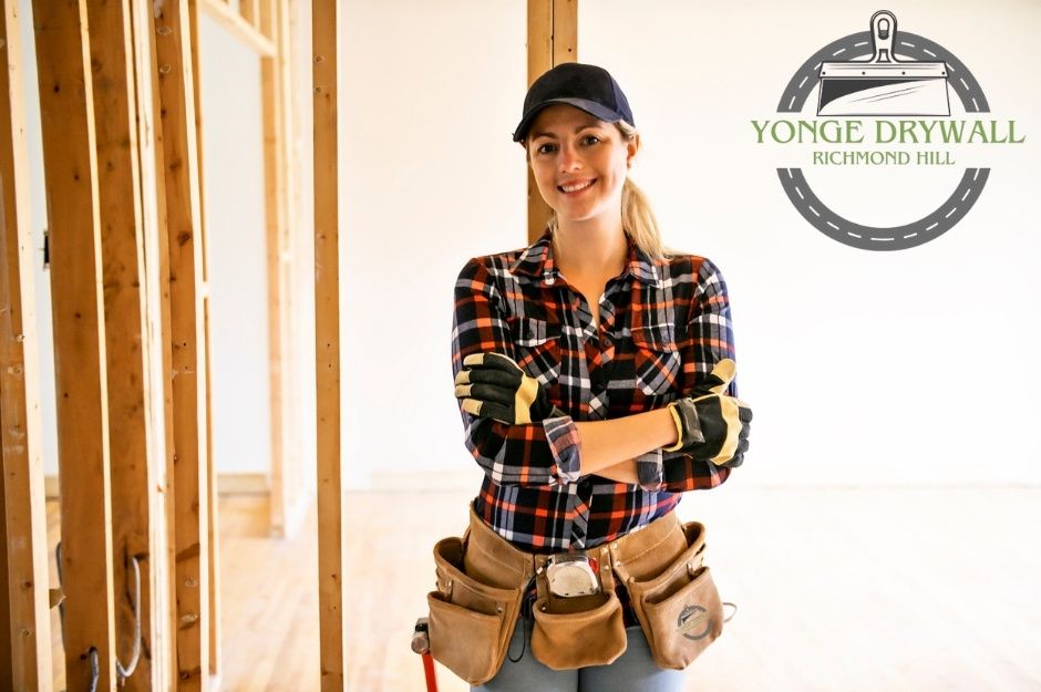 A drywall repair contractor wearing a cap, plaid shirt, and protective gloves stands confidently with arms crossed inside a commercial building under renovation. She has a tool belt filled with tools around her waist. Located near Taunton Rd, and Oak Walk Dr, Oakville, ON.