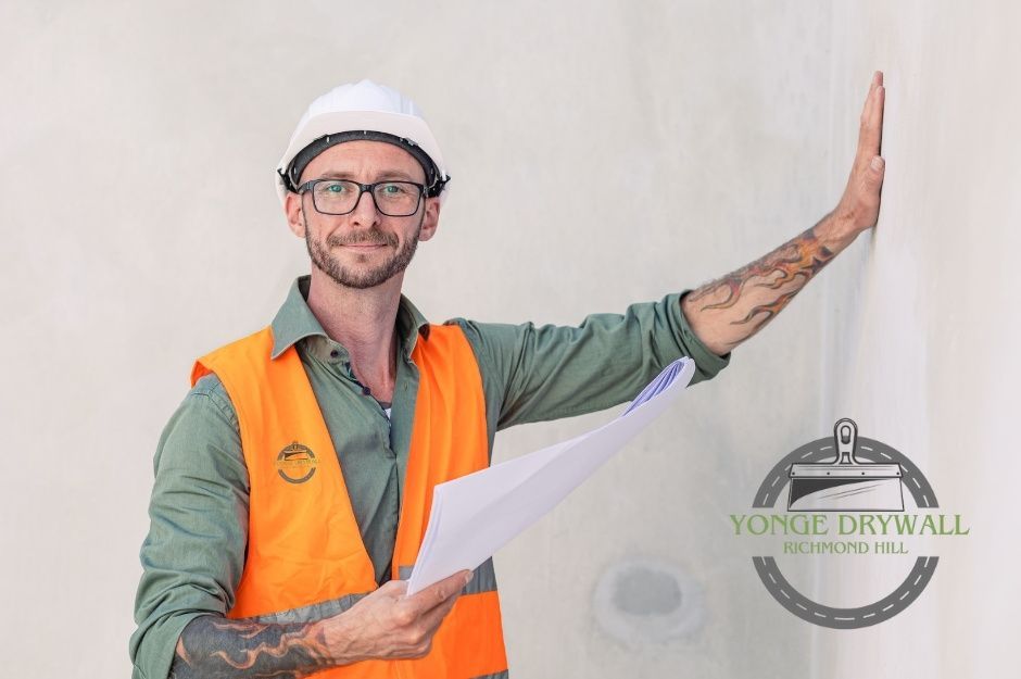 A drywall repair contractor wearing an orange safety vest, green shirt, and white hard hat stands indoors at a residential project, holding blueprints in one hand and inspecting a smooth drywall surface with the other. Located near Central St, and Lorn St, Pickering, ON.