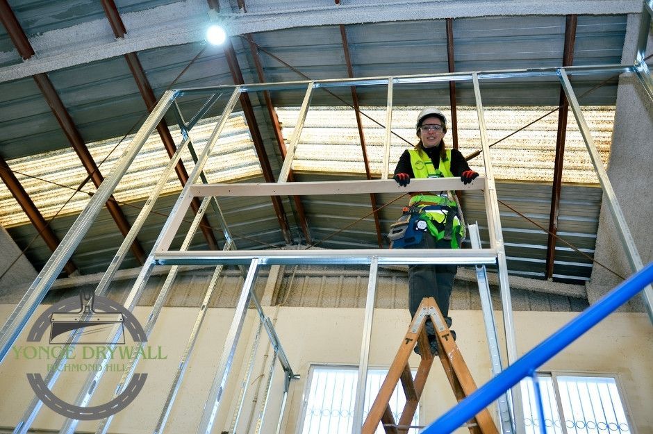 A drywall repair contractor wearing a yellow safety vest, hard hat, and gloves stands on a wooden ladder assembling a metal stud frame for a residential wall. She holds a wooden beam in place, surrounded by exposed steel studs under a high metal roof near Chopin Blvd, and Bathurst Glen Dr, Vaughan, ON.
