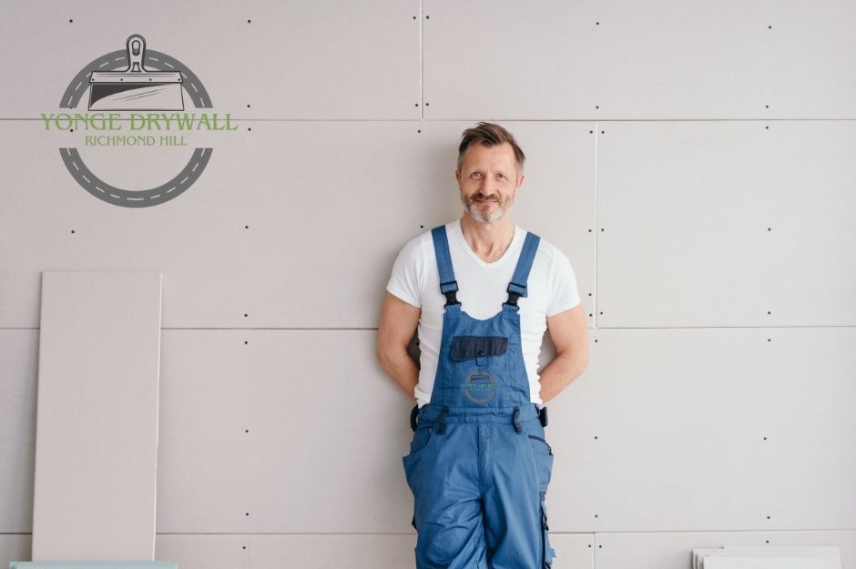 A drywall repair contractor stands in front of a newly installed drywall panel in a commercial project. He is smiling, wearing a white T-shirt and blue overalls with his hands behind his back. Stacked drywall sheets are visible beside him. Located near Ringwood Dr, and Main St, Whitchurch-Stouffville, ON.