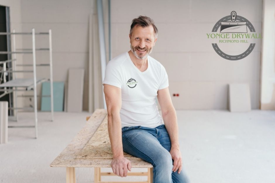 A drywall repair handyman wearing a white t-shirt and jeans is sitting on a wooden workbench in a partially finished room. The background shows drywall sheets, scaffolding, and construction materials neatly arranged, suggesting ongoing residential renovation near Ridge Rd, and Ridge Hill Dr, Aurora, ON.