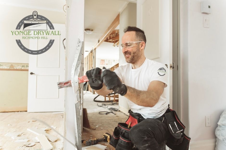 A drywall repair handyman wearing safety goggles, black gloves, and a white t-shirt uses a red crowbar to remove damaged drywall inside a commercial site under renovation. Broken plaster and tools lie on the floor as he works with focus. Located near MacKenzie Dr, and Wilson Dr, Milton, ON.