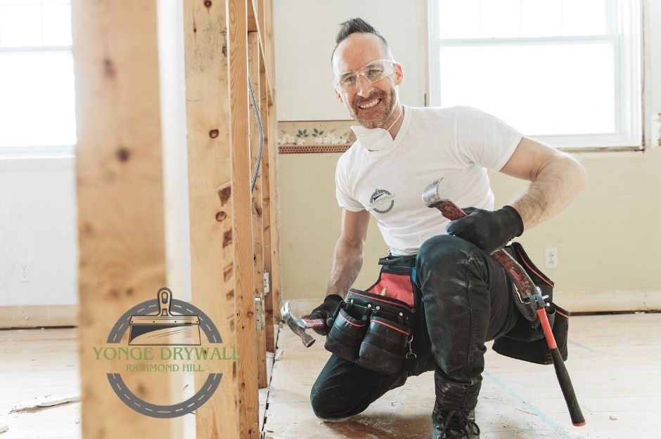 A drywall repair handyman wearing safety goggles, black gloves, and a white t-shirt kneels on a wooden floor holding a hammer in one hand and a crowbar in the other. He smiles during a residential renovation surrounded by exposed wall studs. Located near Foxcroft Blvd, and Quantra Crescent, Newmarket, ON.