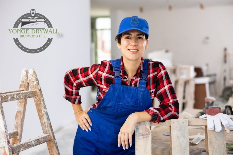 A drywall repair handyman stands confidently in a room under renovation, wearing blue overalls, a red plaid shirt, and a blue cap. She leans on a wooden sawhorse beside a paint-splattered ladder, surrounded by tools in a residential project near Leithridge Crescent, and Carnwith Dr W, Whitby, ON.