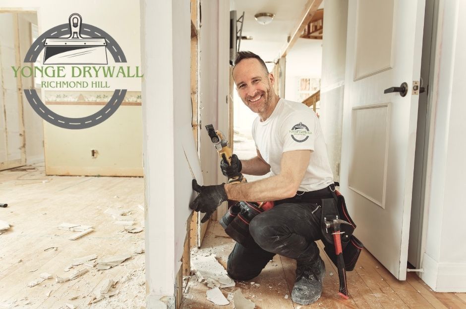 A drywall repair specialist kneels on a dusty floor, smiling while using an oscillating multi-tool to cut damaged drywall during commercial renovation. He wears a white T-shirt, black gloves, and a tool belt with a hammer. Renovation debris surrounds him. Located near Fontaine Dr, and Chartwell Crescent, Georgina, ON.