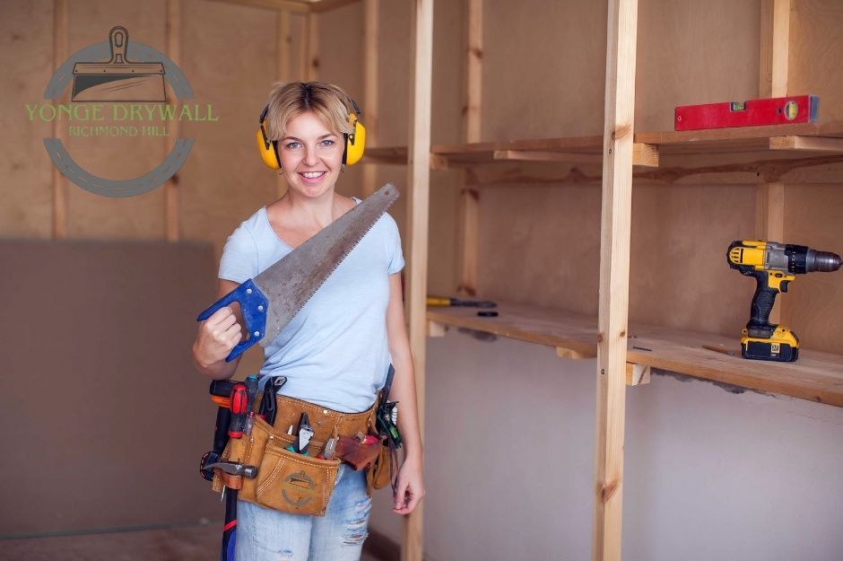 A drywall repair specialist wearing yellow ear protectors and a tool belt full of construction tools stands inside a partially finished commercial room. She smiles while holding a large handsaw with a blue handle, and a yellow power drill rests on the wooden shelf beside her near Reach St, and N Port Rd, Scugog, ON.