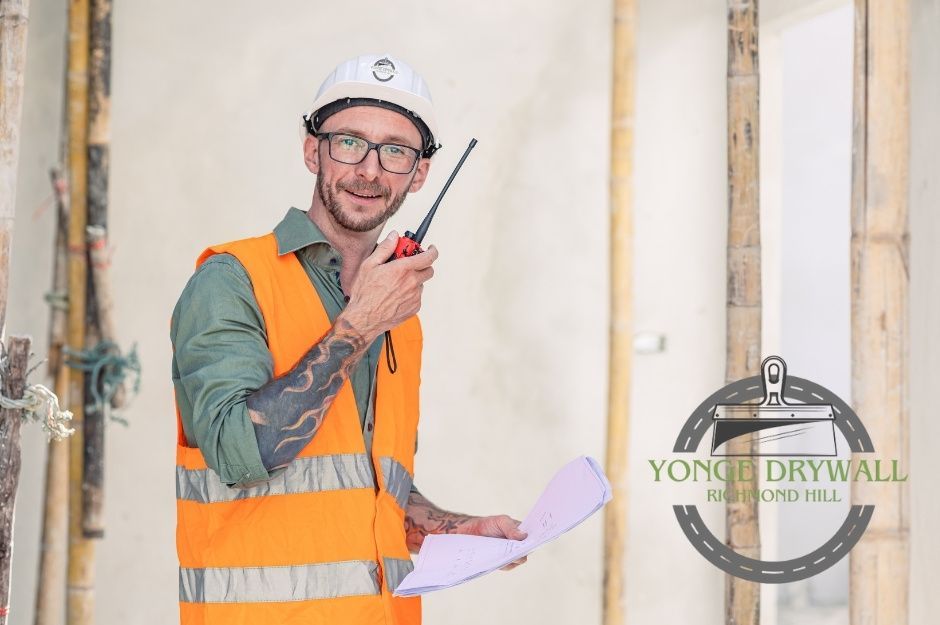 A drywall repair technician stands in a commercial project space, wearing an orange safety vest with reflective stripes, a white hard hat, and glasses. He holds a two-way radio in one hand and a set of blueprints in the other. Located near Cornwall Dr, and Coles Ave, Ajax, ON.
