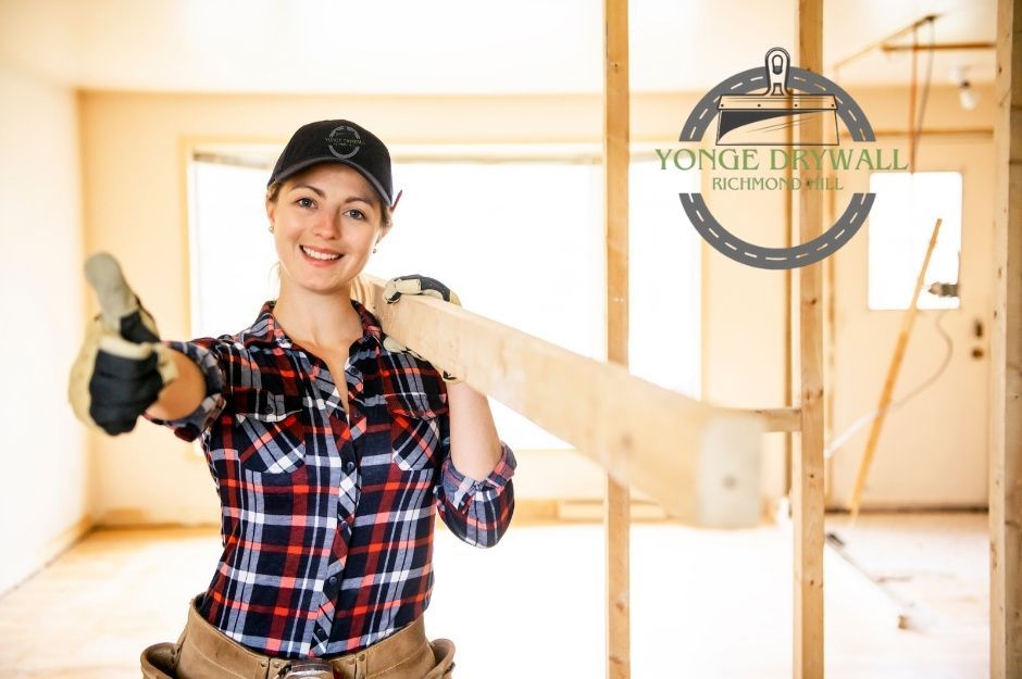 A drywall repair technician in a plaid shirt and black cap carries lumber on her shoulder in a residential construction site. The workspace shows ongoing residential construction and framing work near Joseph St, and Main St, Halton Hills, ON.