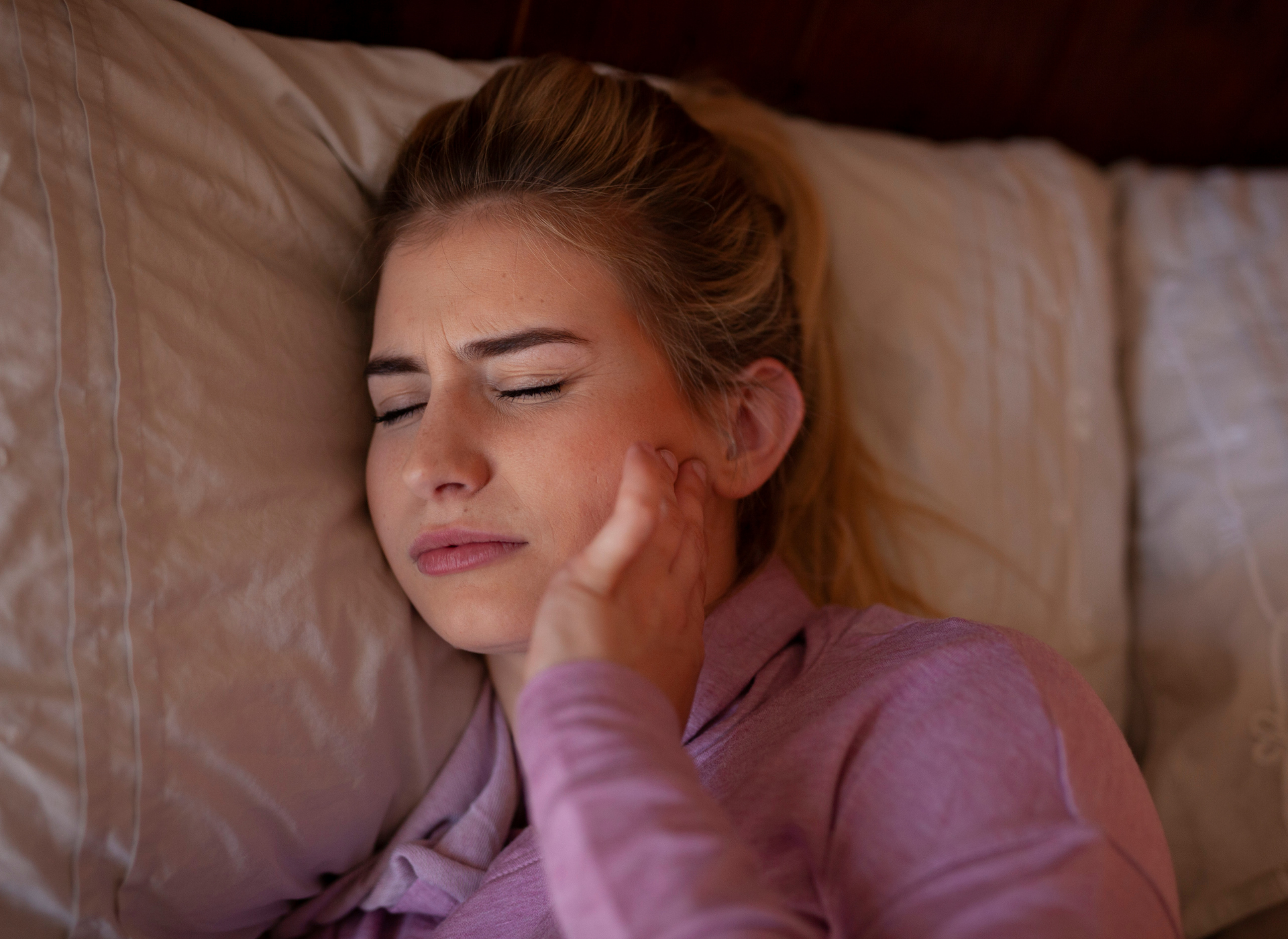Woman lying in bed, holding her jaw, appearing to be in pain.