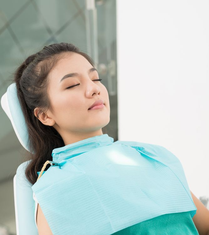 Woman in dental chair, eyes closed, wearing a bib, under bright light.