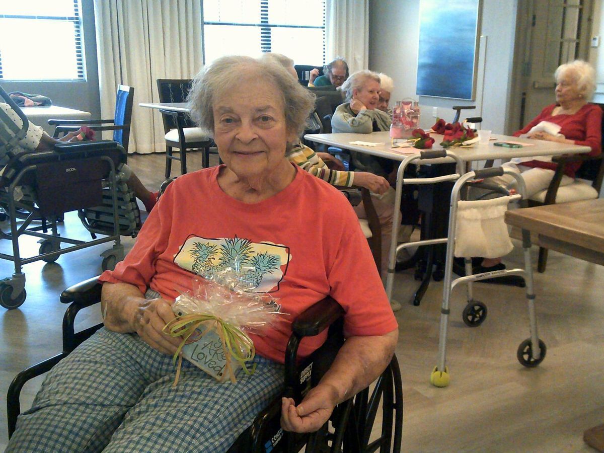 Elderly woman in wheelchair smiling, holding a gift, in a community room with other seniors.