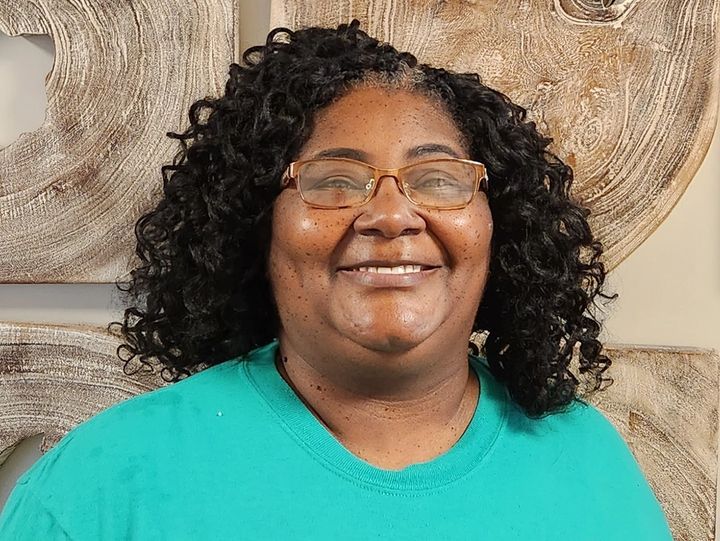 Woman with curly black hair and glasses smiles, wearing a teal shirt, against a textured wall.