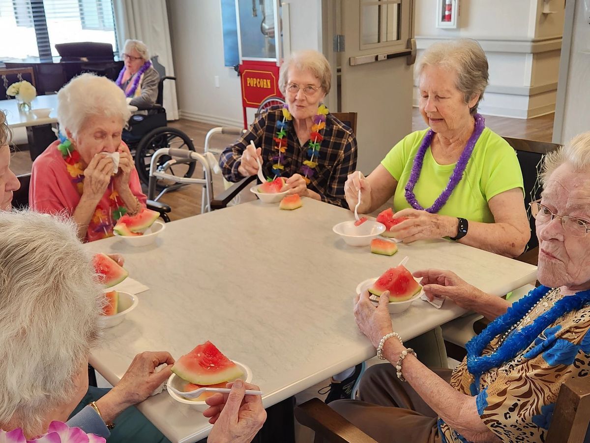 A group of elderly women enjoy watermelon, wearing leis, seated around a table.