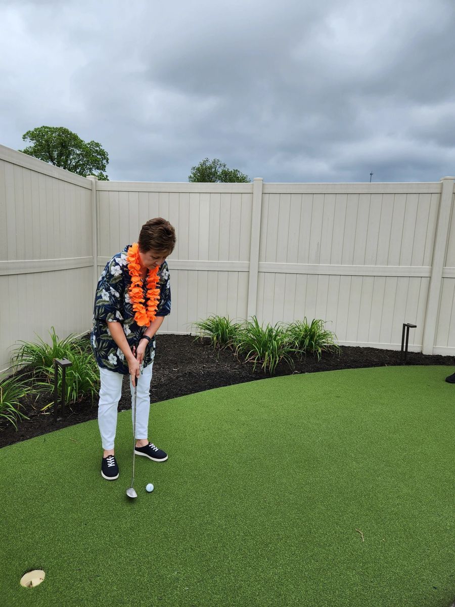 Person putting a golf ball on a green, wearing a floral shirt and lei, against a white fence.