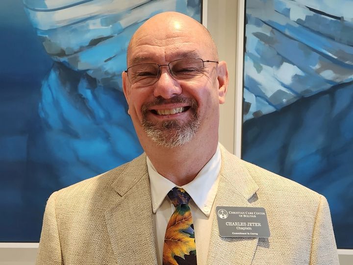 Man with glasses and a patterned tie smiles, wearing a blazer, standing in front of a blue artwork.