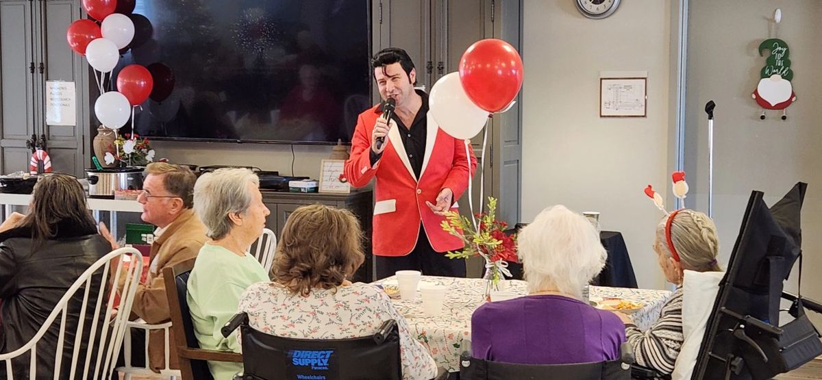 Elvis impersonator singing to seniors at a holiday party, red and white balloons, decorations.