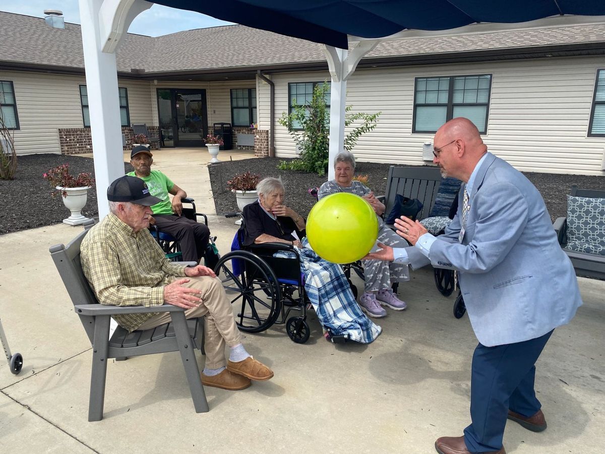 People playing with a yellow ball outside. Elderly residents and a man in a blazer enjoy a game under a canopy.