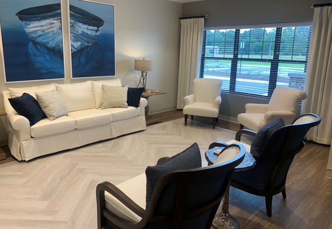 Living room with white sofa, blue boat art, white chairs, and large window overlooking a yard.