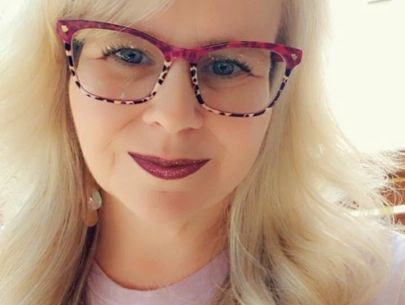 Woman with blonde hair and pink leopard-print glasses smiles, dark lipstick.
