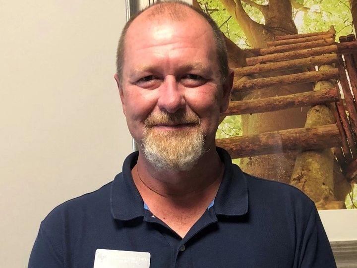 Man with a short beard in a blue shirt, smiling at the camera. Background has a treehouse picture.