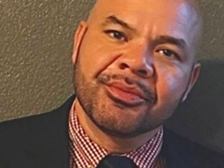 Bald man, slight smile, wearing a dark suit jacket, and patterned red and white shirt.