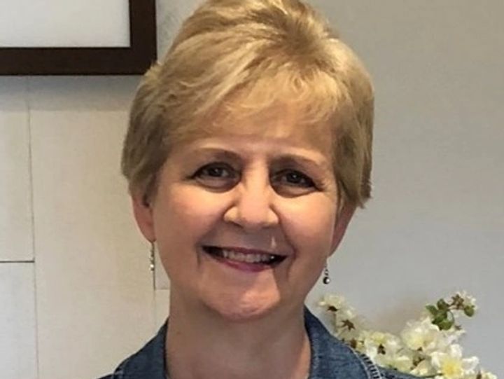 Blonde woman smiles; wearing a denim jacket and earrings, in front of a white wall with flowers.