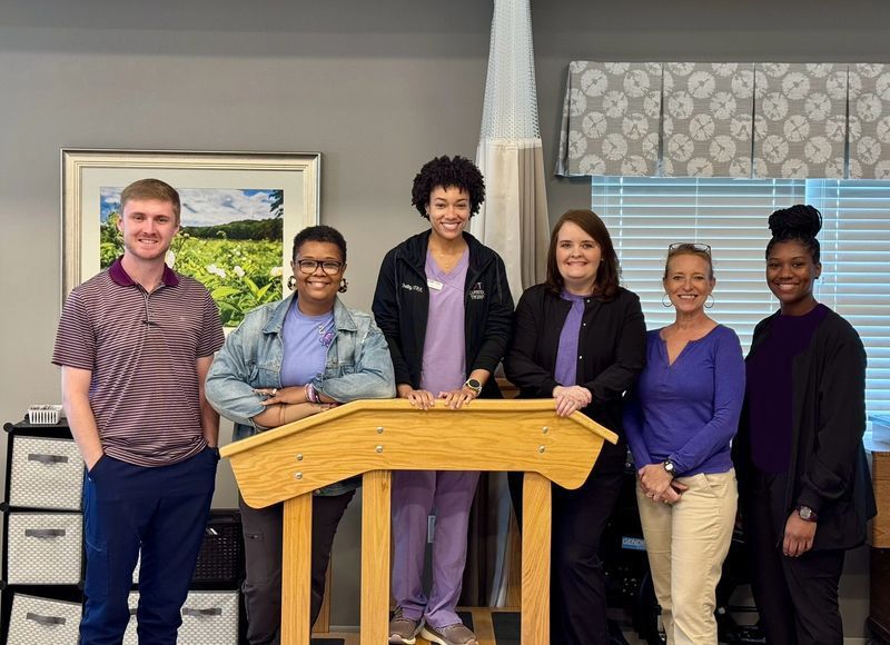 Group of six healthcare workers posing, smiling by wooden equipment.