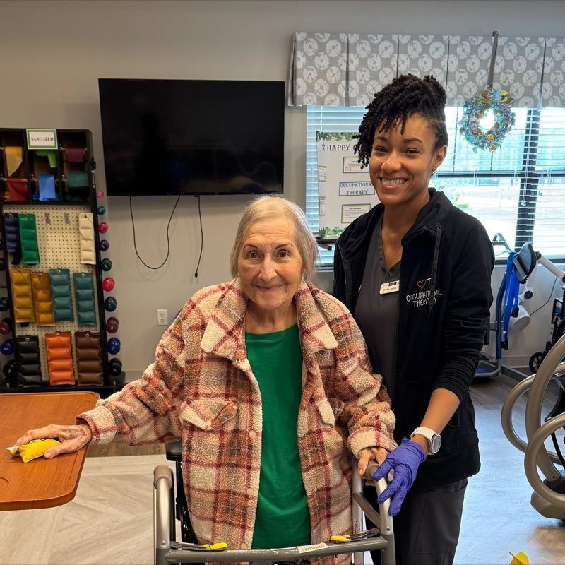 An elderly woman in a walker and a younger caregiver smile in a rehab center.