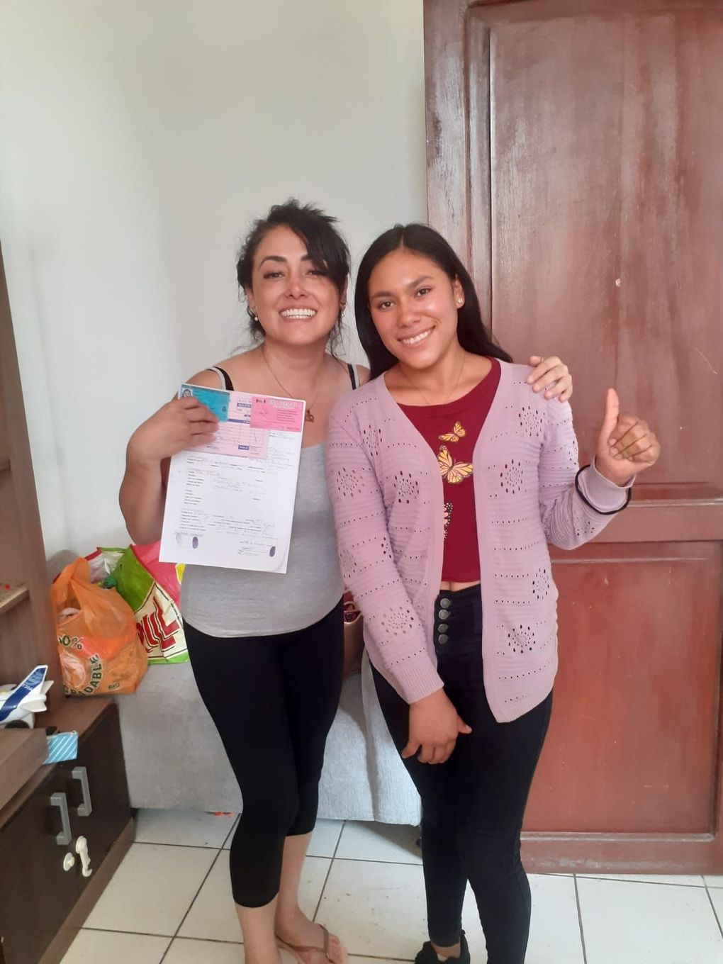 Dos personas sonrientes posan en el interior de una puerta de madera; una sostiene un periódico y la otra levanta el pulgar.