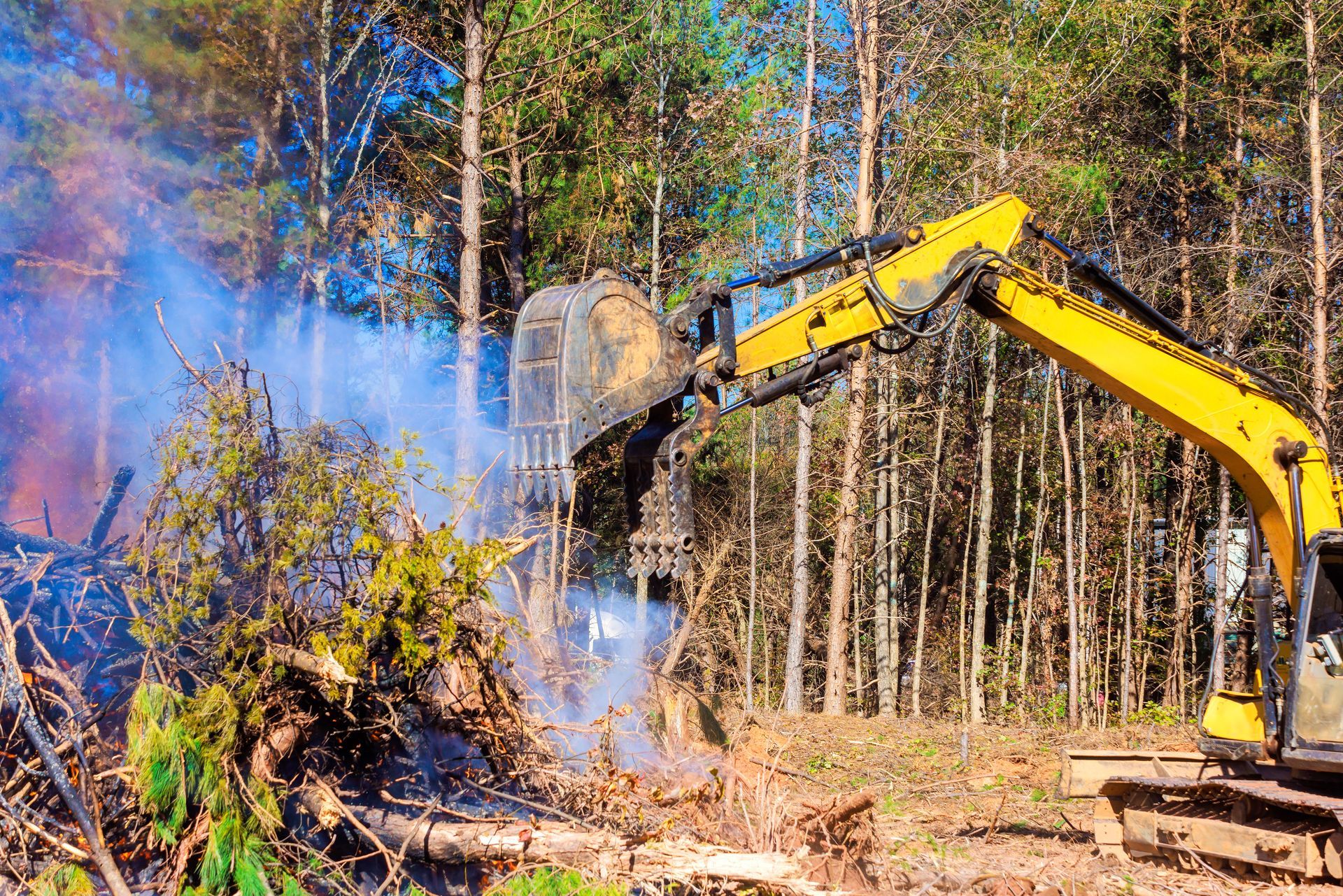 Excavator clearing brush in a forest, with blue smoke rising near cut trees