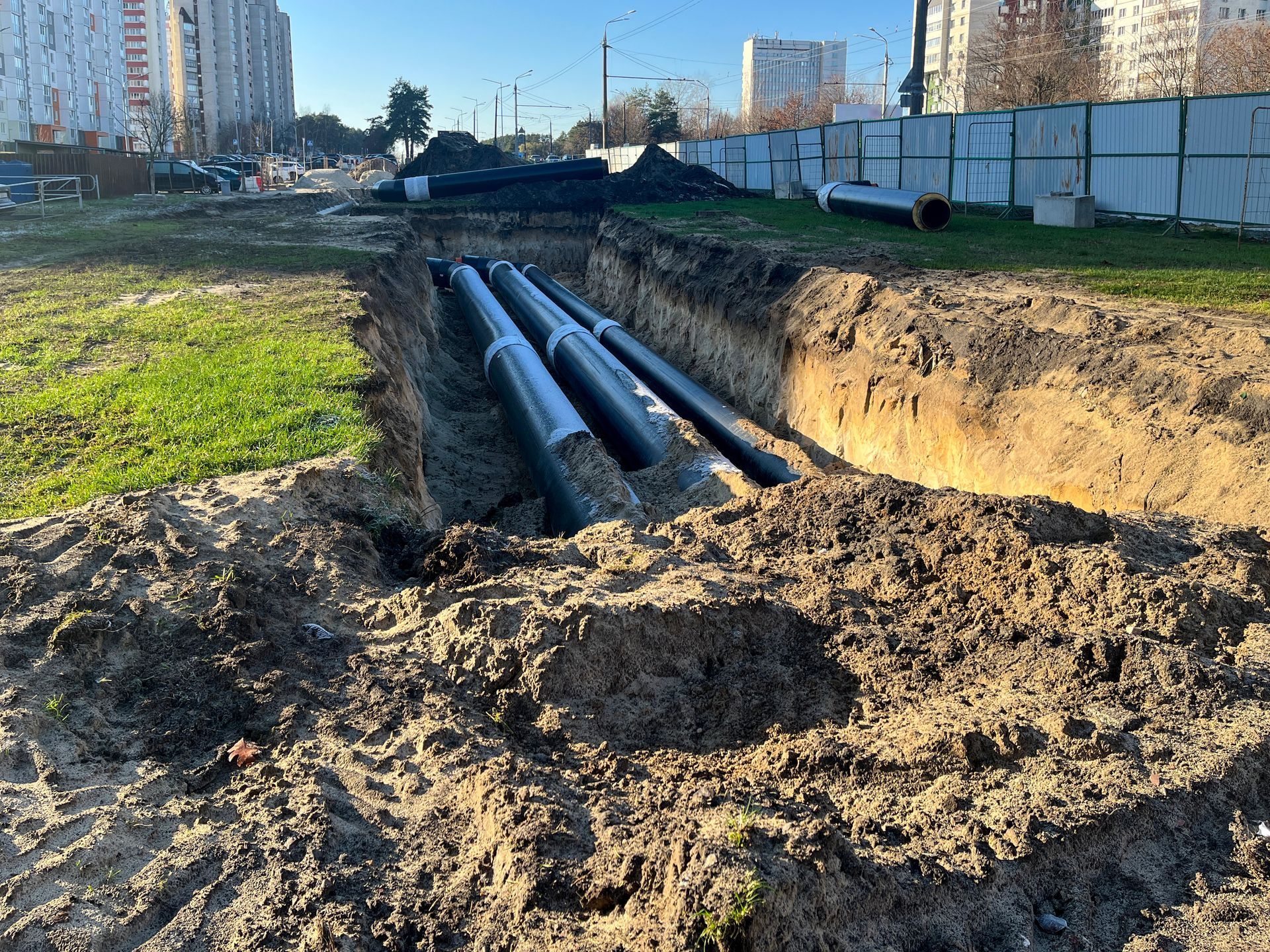Trenches with large underground pipes beside a muddy construction site in a city neighborhood