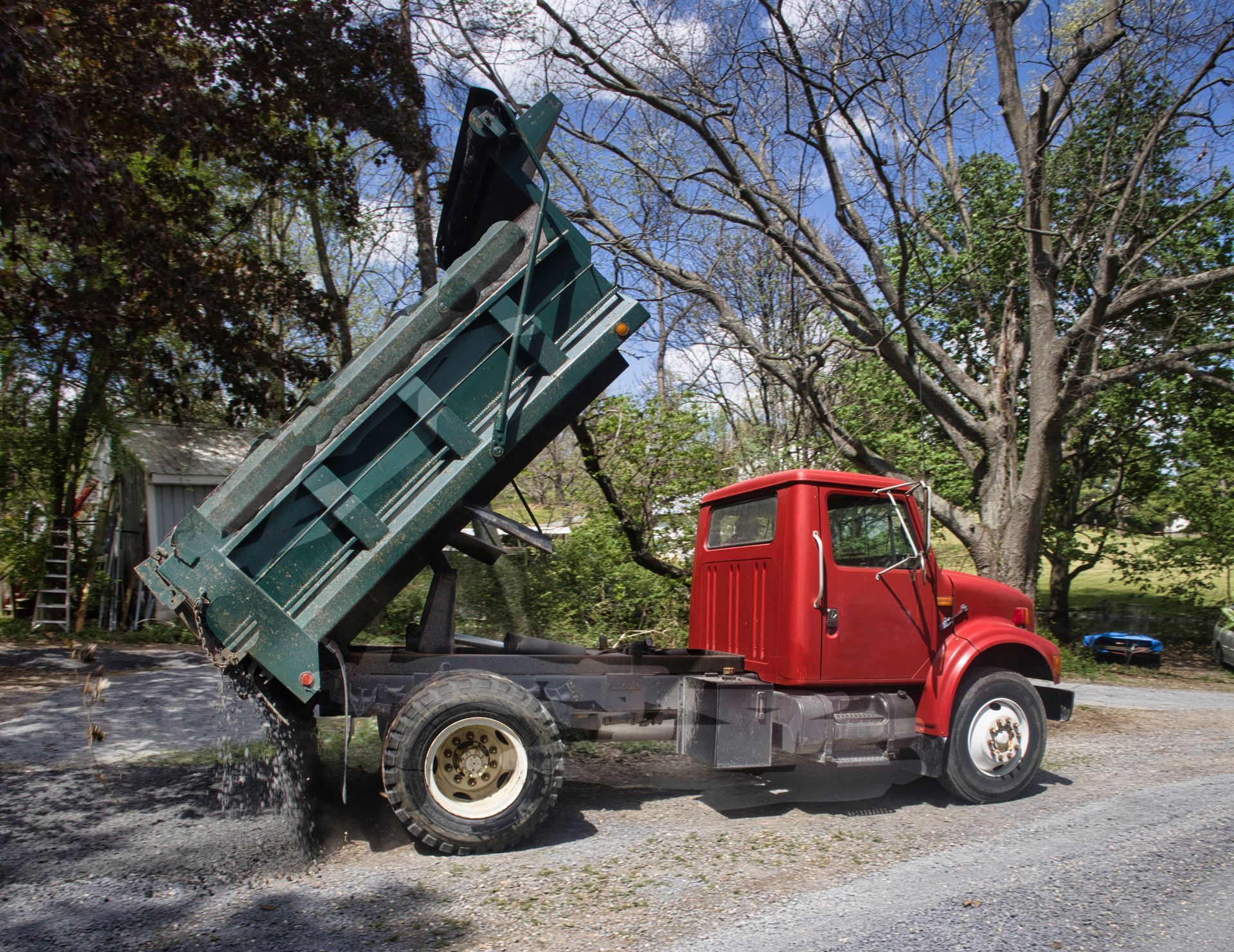 Red dump truck with raised green bed parked on a gravel road beside trees