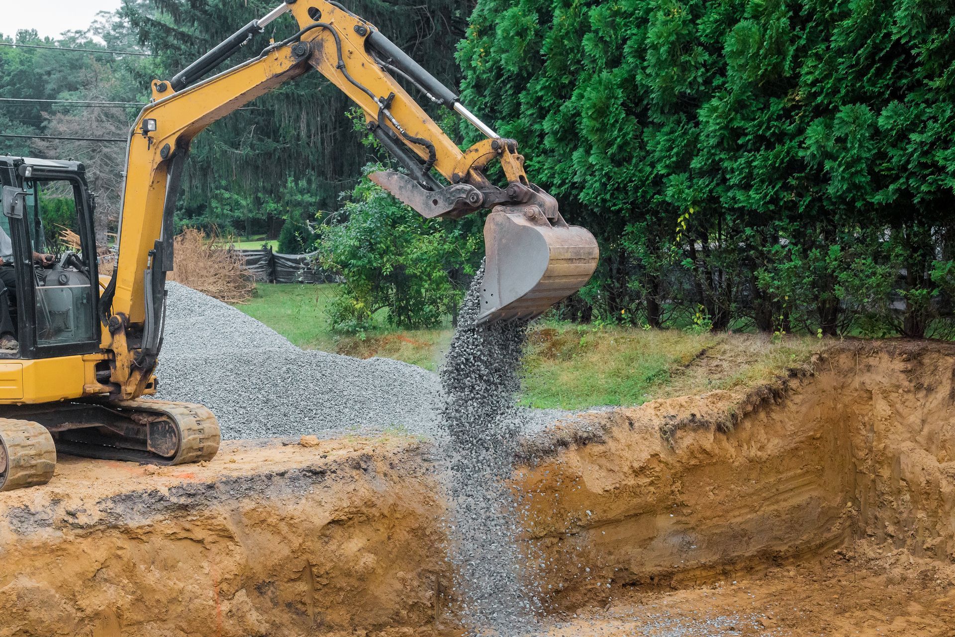 Yellow excavator dumping gravel into a trench at a construction site outdoors