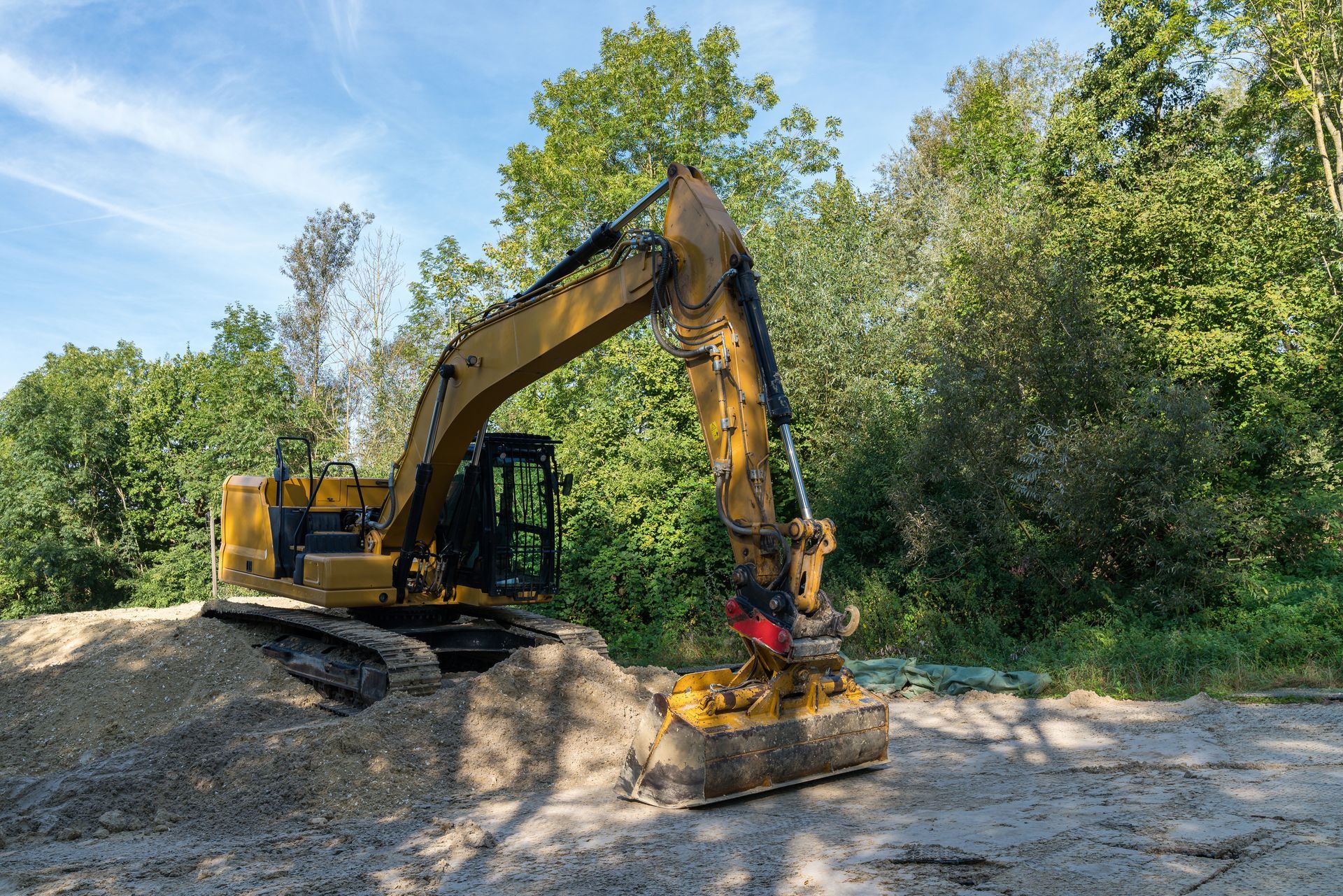 Yellow excavator clearing dirt on a sunny roadside near trees