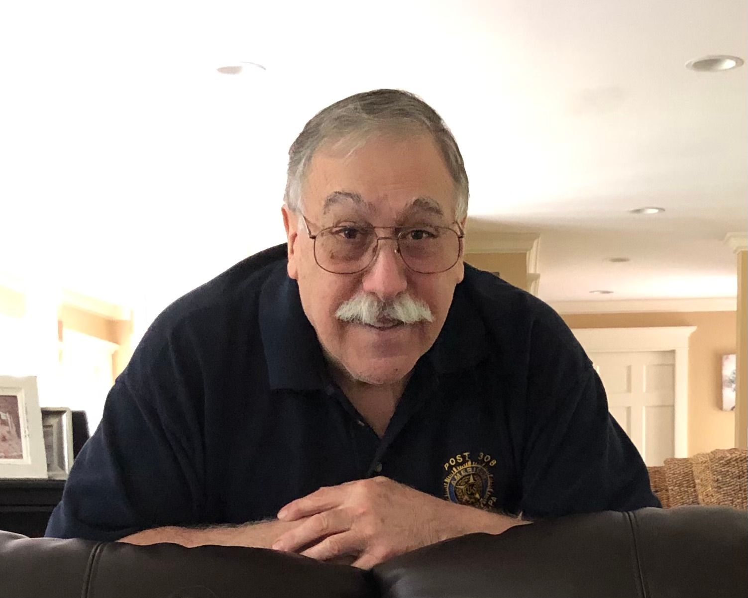 Older man wearing glasses, mustached and a dark blue shirt, smiling at the camera, indoors.