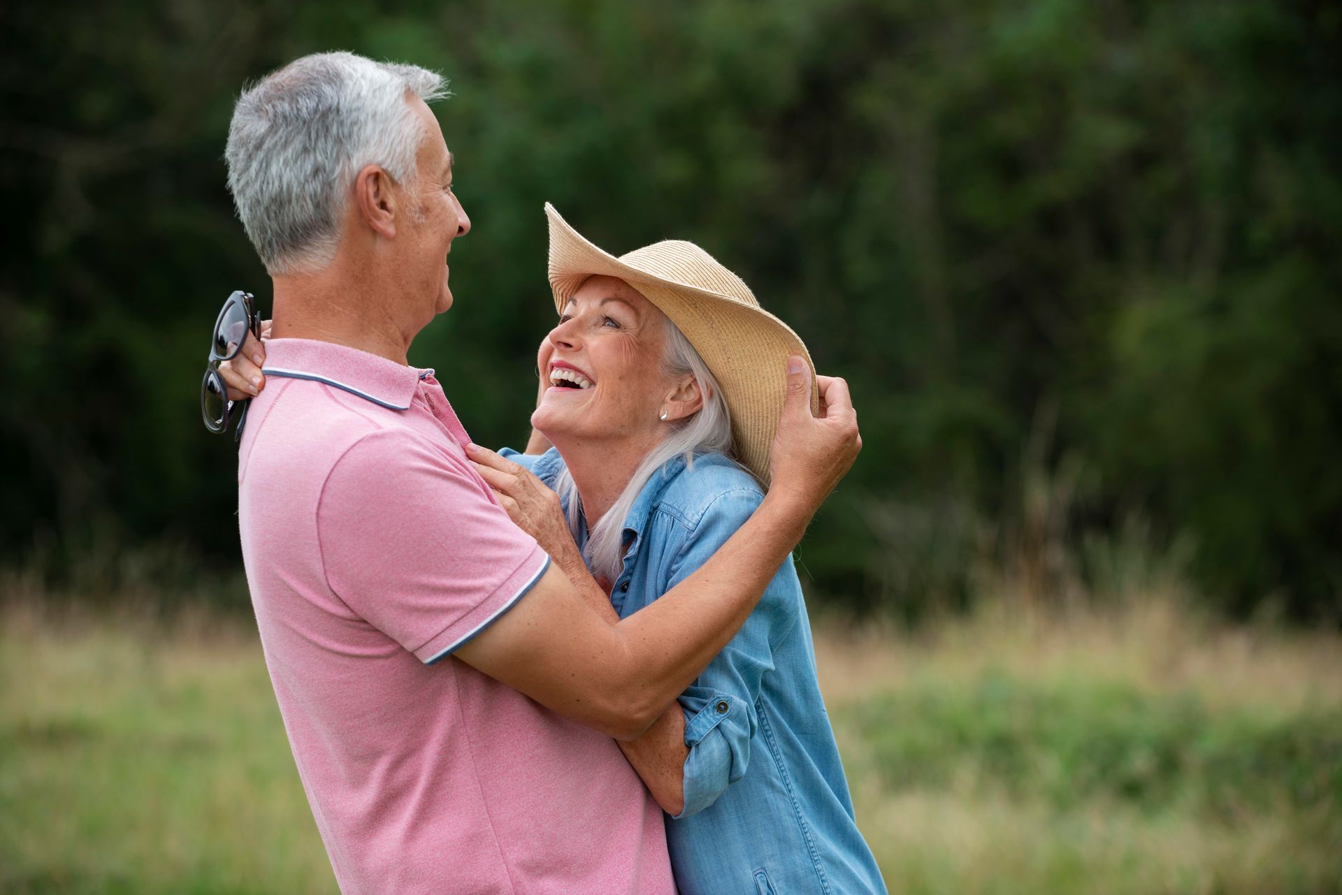Senior couple embracing and laughing in a field. Man in pink shirt, woman in denim, straw hat.