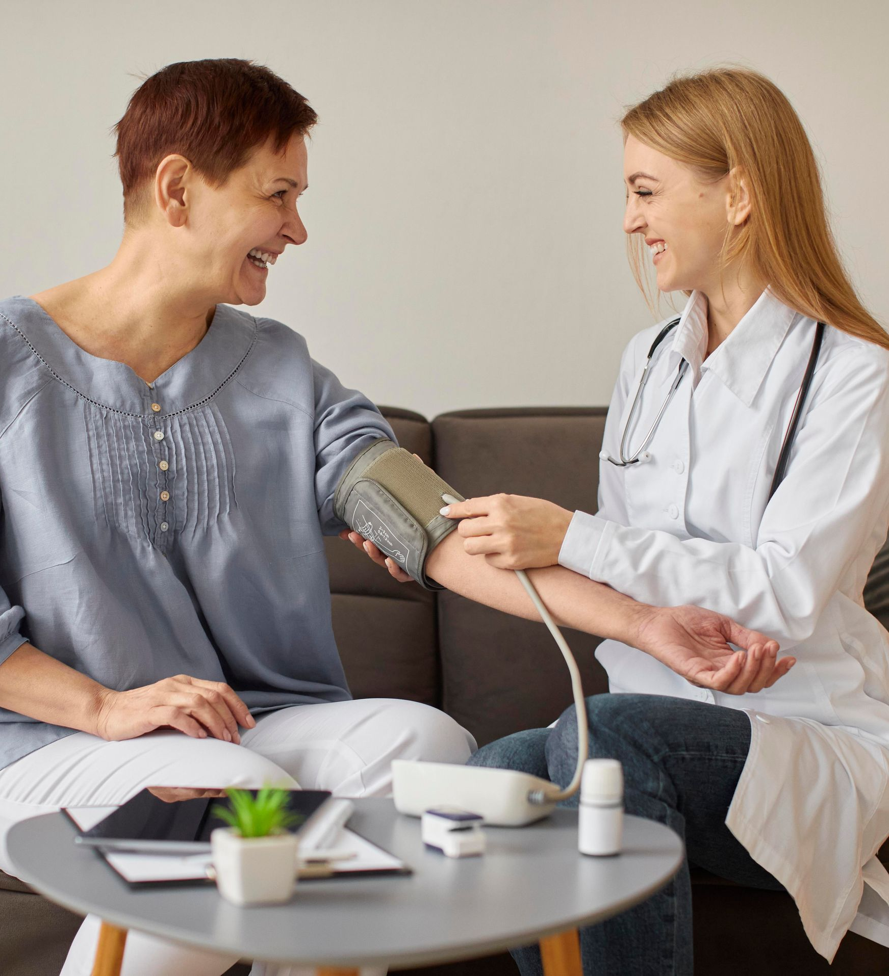 Doctor taking a patient's blood pressure in a home setting. Both are smiling, with the doctor in a white coat.