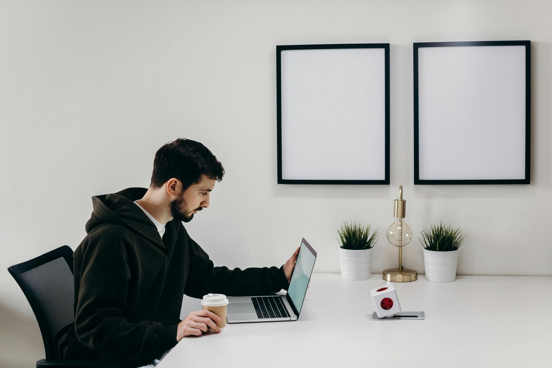 Man in a black hoodie working on a laptop at a desk with coffee and two blank frames on the wall.