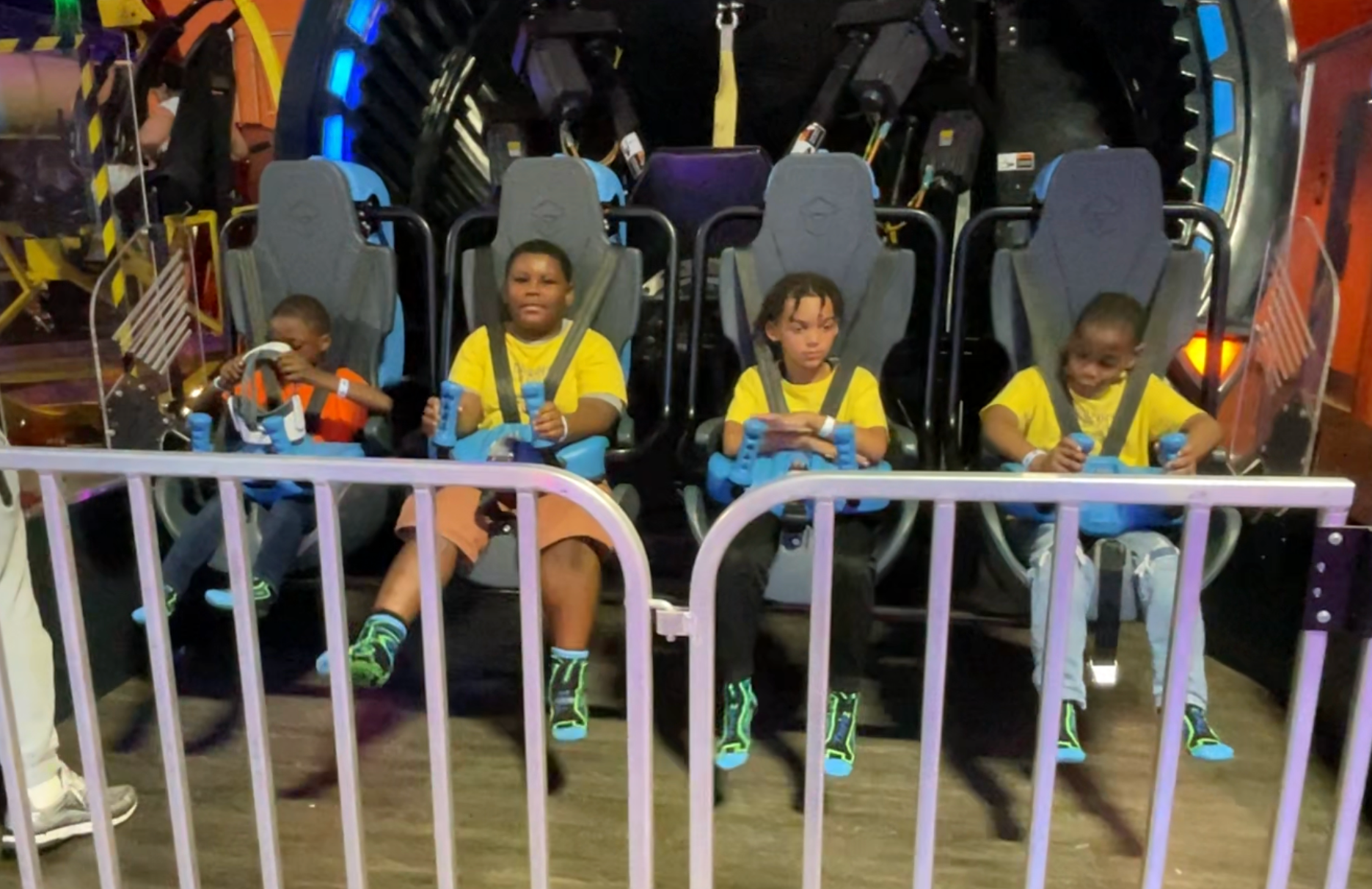 Four children in yellow shirts on a ride, sitting in blue seats, holding on. Amusement park setting.