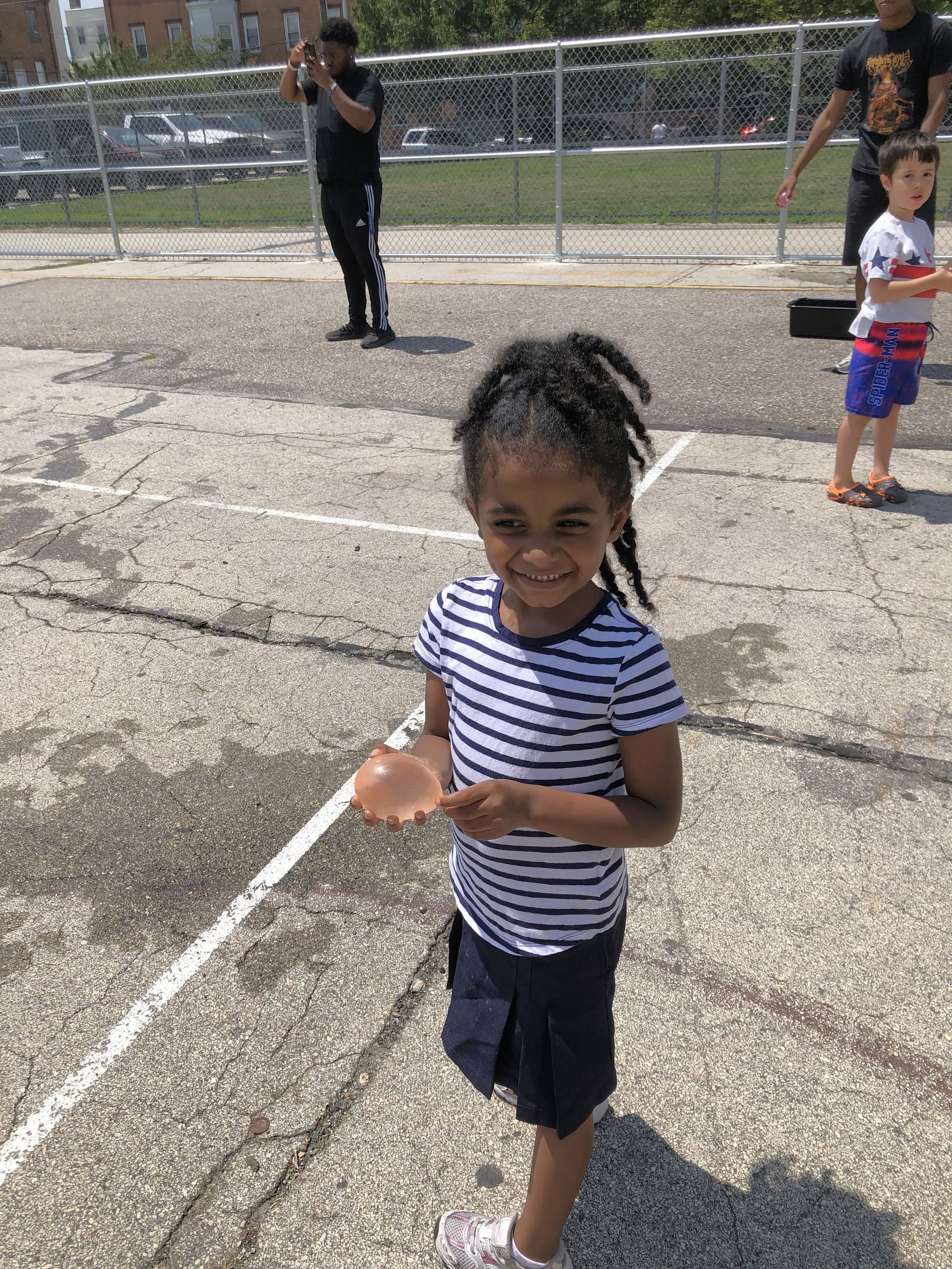 Smiling girl in striped shirt and skirt holds water balloon on a sunny blacktop, other people visible.
