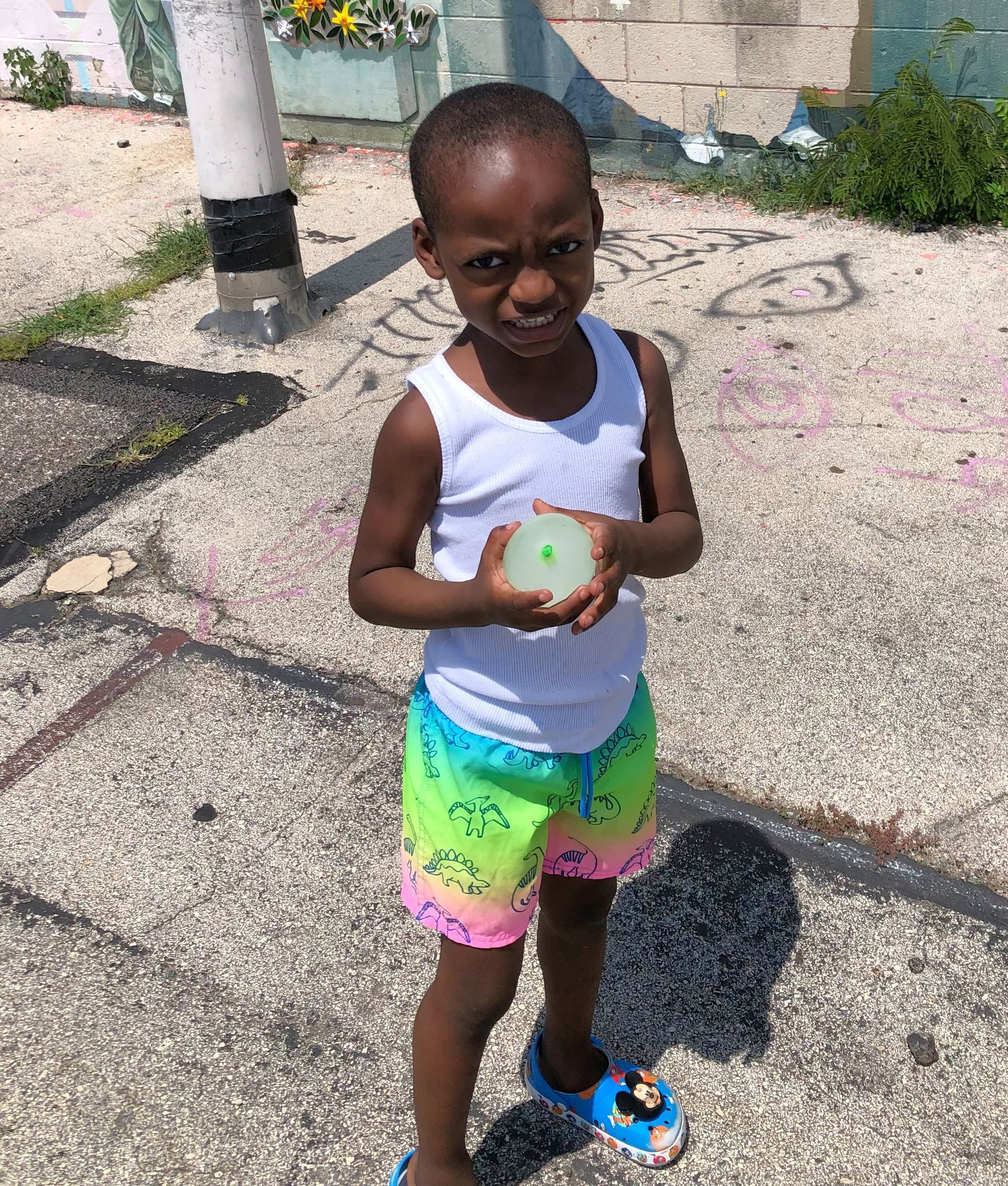Young child with dark skin, holding a translucent ball, smiles on a sidewalk in colorful shorts.