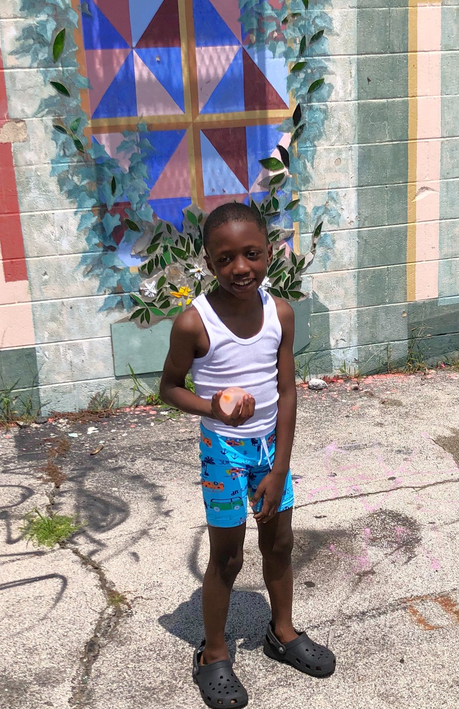 Boy in a white tank top and blue swim trunks, standing in front of a colorful mural.