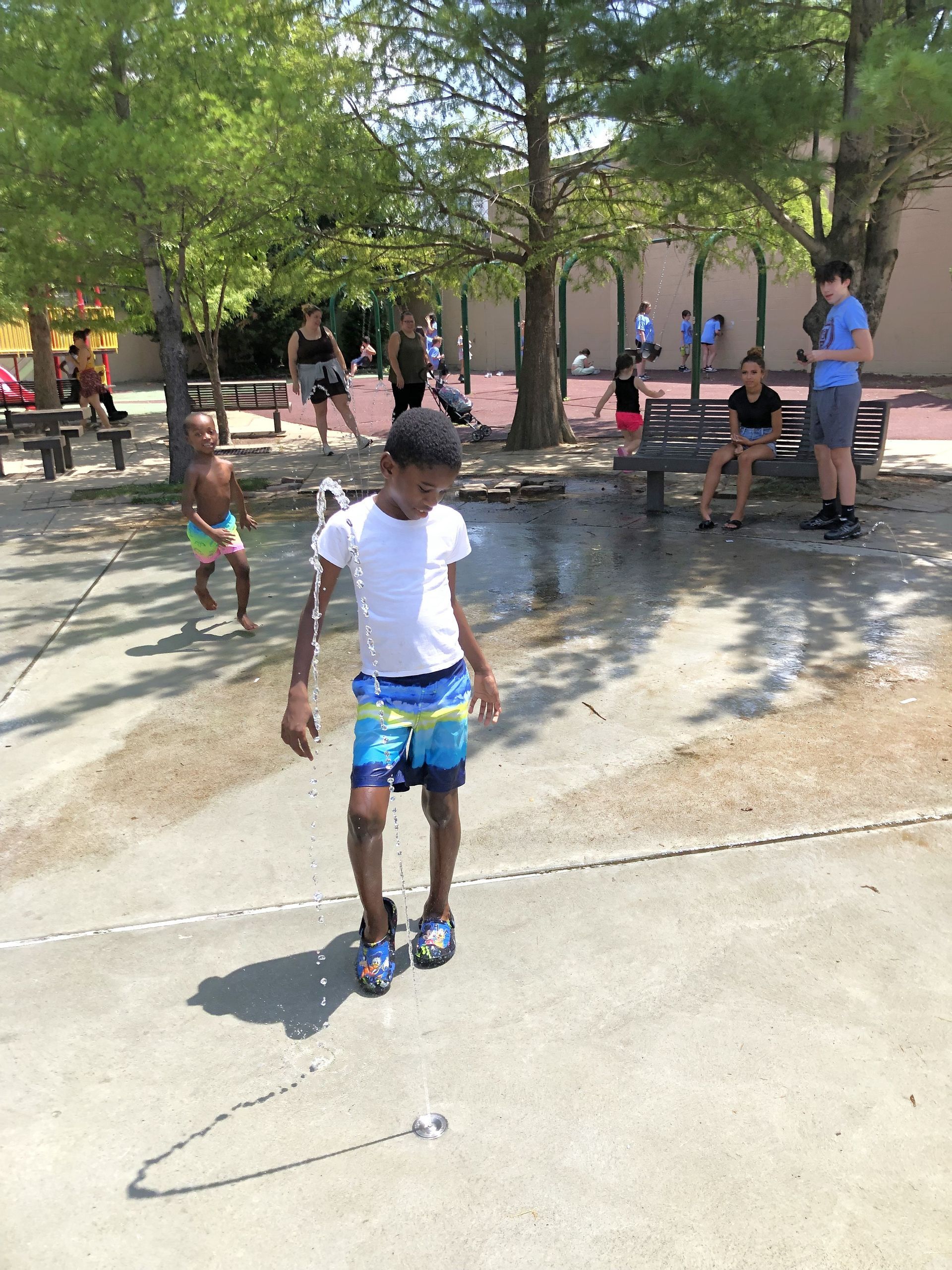 Boy in swimsuit playing in water spray at a park with other people and trees.