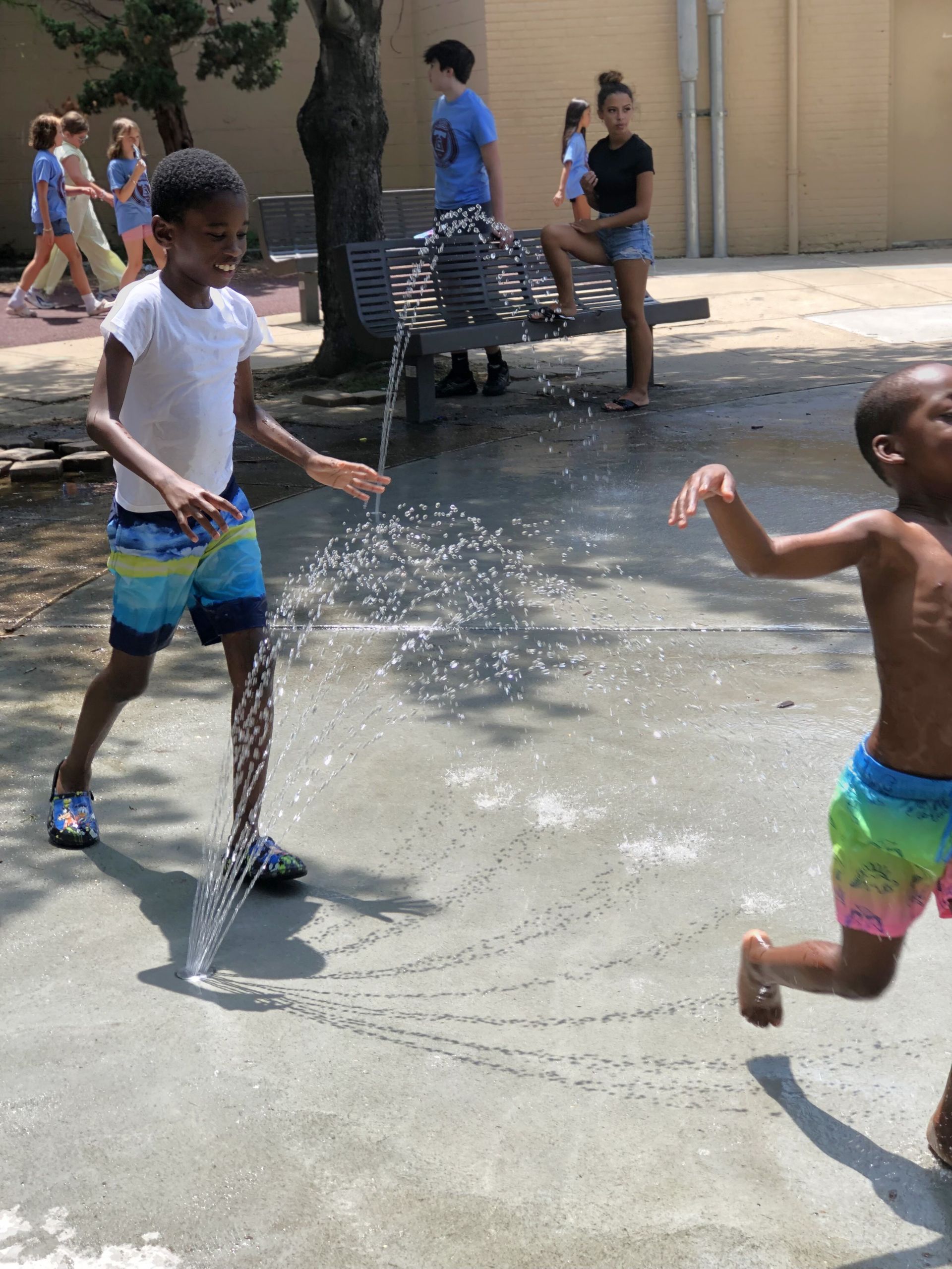 Two Black children play in a water fountain, one splashing the other. Others watch.